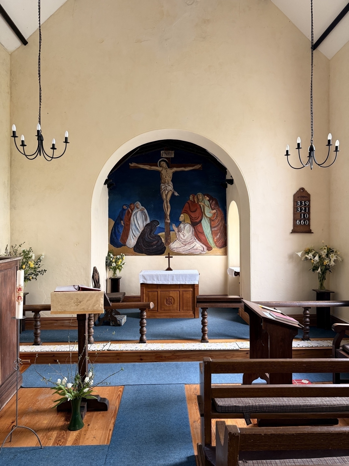 A small church interior features a mural of the Crucifixion above the altar, surrounded by pews and floral arrangements.